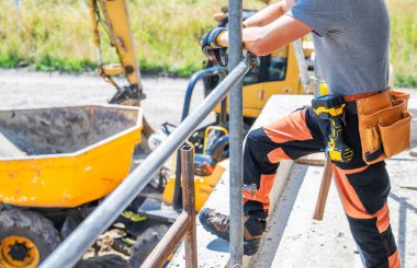 Sarı makineler arka planda açık gökyüzünün altında çalışırken, bir inşaat işçisi görev yapmak için elektrikli aletler kullanarak bir çerçevenin üzerinde duruyor..