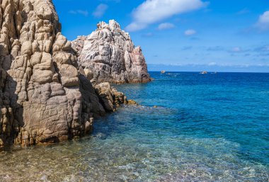 Rocky cliffs rise above crystal clear water under a bright blue sky of northern Sardinia Italian Island.