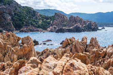Rocky cliffs rise majestically along the northern Sardinia shoreline, with turquoise waves lapping at the stones under a sunny sky in Italian coastal landscape.