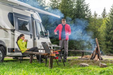 Bir erkek kamp ateşinin üzerinde yemek hazırlarken, bir kadın sakin bir ormanda karavanın yanında oturmuş sıcak bir içkinin tadını çıkarıyor..
