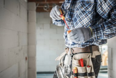 Yetenekli bir elektrikçi eldiven giyerken aletler kullanarak bir lamba tesisatı kurmaya çalışıyor. İnşaat alanı kısmen bitmiş ve iyi aydınlatılmış..