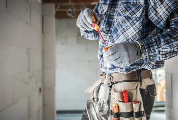 Yetenekli bir elektrikçi eldiven giyerken aletler kullanarak bir lamba tesisatı kurmaya çalışıyor. İnşaat alanı kısmen bitmiş ve iyi aydınlatılmış..