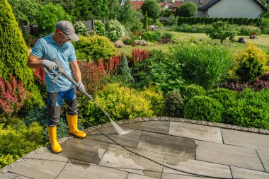 Sarı çizmeli bir adam çiçek ve bitkilerle dolu canlı bir bahçedeki taş bir verandada basınç makinası kullanıyor..