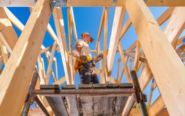 Bir inşaat işçisi, güneşli bir günde inşaat projesine tahta kirişleri yerleştirirken iskelenin üzerinde durur..