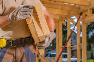 Bir inşaat işçisi gün ışığında kısmen inşa edilmiş bir binanın içinde dururken ahşap kalasları tutar..