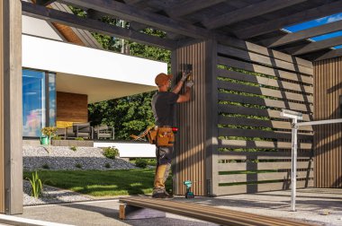 Bir işçi gün boyunca arka bahçeye ahşap paneller kuruyor. Ev modern tasarıma ve yeşil manzaraya sahip..