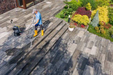 Bir işçi bahçede taş bir yüzeyi temizlemek için basınç makinesini kullanır. Bölgede bitkiler ve renkli çiçekler var..