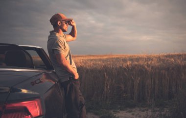 Cabrio marka üstü açık arabasının yanında duran bir adam gün batımında bulutlu bir günde ufka doğru bakıyor..