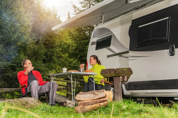 İki kişi bir kampçının yanında bir masada oturur, gün ışığında ormanda yiyecek ve içecekleri paylaşırlar..