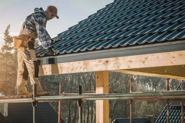 Bir inşaatçı bir eve çatı kiremitleri kurmak için iskelenin üzerinde durur. Güneş berrak bir gökyüzünde parlıyor.