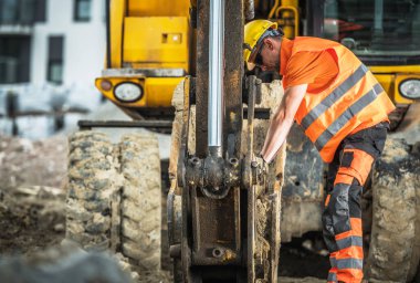 Turuncu gömlekli bir işçi inşaat alanında makine kullanıyor. Olay yeri kir ve makineleri gösteriyor..