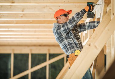 Bir işçi, gün ışığında bir inşaat sahasında tahta kirişleri güvence altına almak için matkap kullanır..