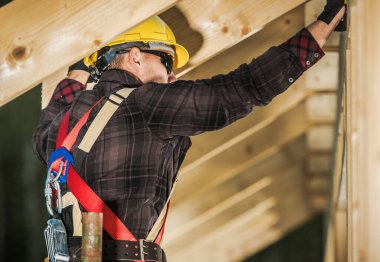 A worker in a yellow helmet fixes wooden beams at a building site in the daylight.