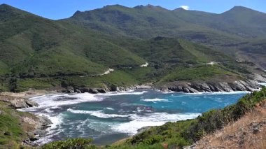 Dalgalar kıyıya vuruyor. Korsika sahilinde. Arka planda tepeler var..