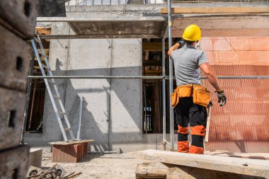 Bir inşaat işçisi, sarı bir başlık ve güvenlik teçhizatı giyerken duvarla ilgili malzemeler üzerinde çalışıyor..