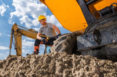 Bir işçi inşaat malzemelerinin yanında bulutlu açık bir gökyüzünün altında toprağı kazıyor..