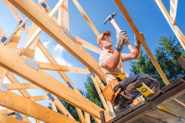 Bir işçi inşaat alanındaki tahta kirişleri korumak için çekiç kullanır. Gökyüzü açık ve parlak.