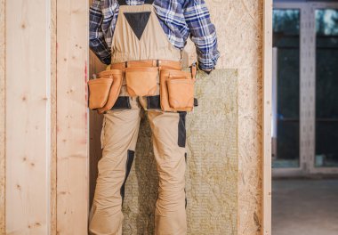 Bir işçi, ahşap bir duvara izolasyon yerleştirirken inşaat alanında duruyor..