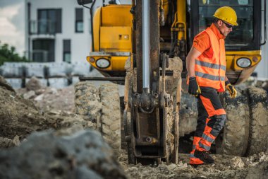Parlak teçhizatlı bir inşaat işçisi gündüz ağır makinelerin yanında yürür..