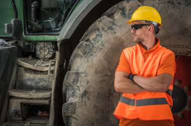 Güvenlik donanımlı işçiler, şantiyede büyük bir traktörün yanında dikilirken dışarıya bakarlar..