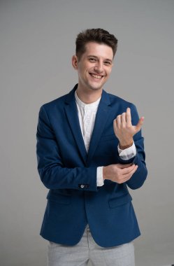 Genç bir adam parlak mavi bir ceketle kelepçesini düzeltirken parlak bir şekilde gülümsüyor. Modern, minimalist bir stüdyoda kendine güveni tam bir şekilde duruyor..