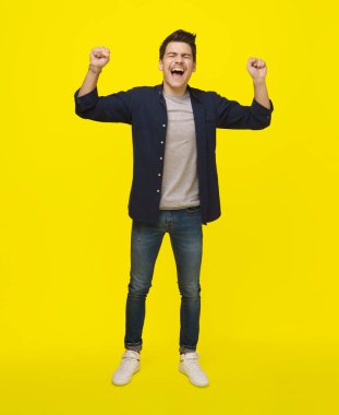 Donanma gömleği ve kotlu genç bir adam parlak sarı bir zemine karşı duruyor, kolları havada ve ağzı açık muzaffer bir haykırışla.