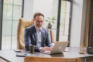 Kendini işine adamış profesyonel bir adam şık ofis masasında verimli bir iş gününün tadını çıkarır. Dizüstü bilgisayarıyla tamamen meşguldür. Görevleri sırasında neşe ve konsantrasyon yayar..