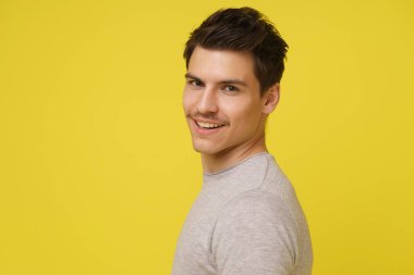 Kısa kahverengi saçlı ve açık gri gömlekli genç bir adam parlak sarı bir arka plana karşı duruyor, başını hafifçe yana çevirmiş, kameraya bakarken gözleri neşeyle parıldıyor..