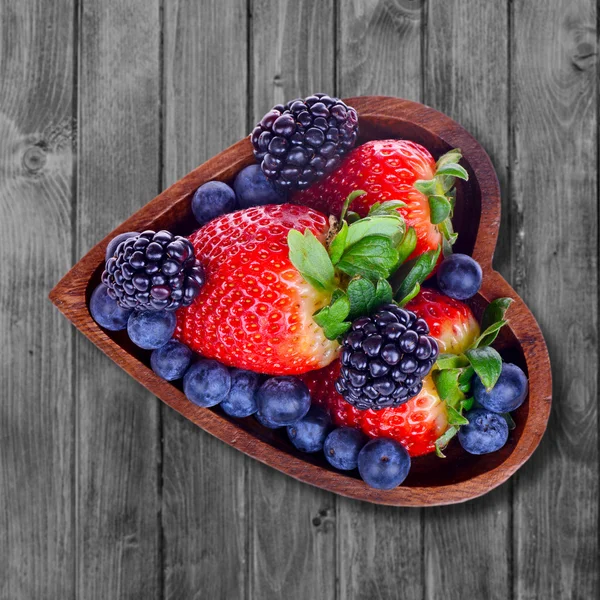 Various berries in a bowl in the form of heart on an old wooden surface — Stock Photo, Image