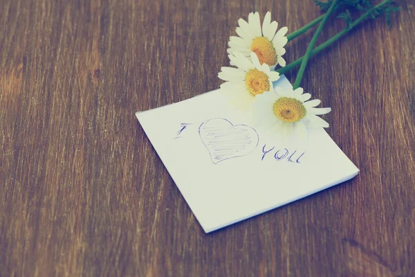 Daisy flower and piece of paper with text "I love you" on the wooden table. Photo in vintage style — Stock Photo, Image