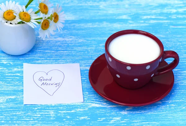 Daisy bloemen en kopje melk met opmerking Goedemorgen op rustieke blauwe houten achtergrond — Stockfoto