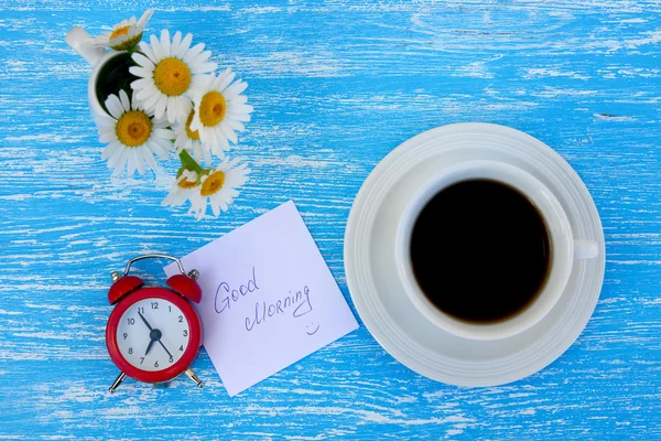 Madeliefjebloemen, wekker en kopje koffie met opmerking Goedemorgen op rustieke blauwe houten achtergrond — Stockfoto