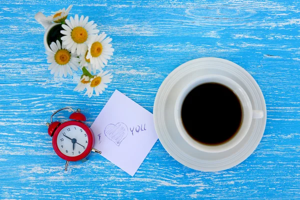 Madeliefjebloemen, wekker en kopje koffie met ik hou van je opmerking op rustieke blauwe houten achtergrond — Stockfoto
