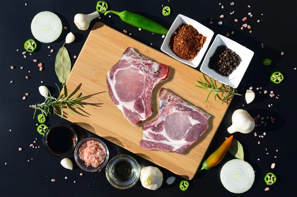 Carne di maiale cruda su un tagliere e pepe, foglia d'alloro, rosmarino, cipolle, sale, olio d'oliva, salsa su uno sfondo nero — Foto Stock