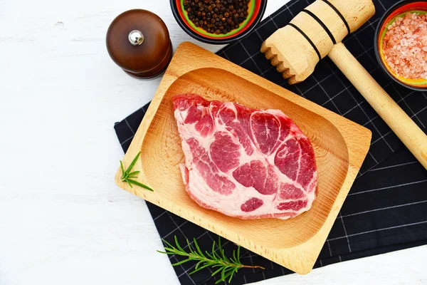 Carne di maiale cruda su piatto di legno, rosmarino, spezie e martello su fondo bianco e nero — Foto Stock