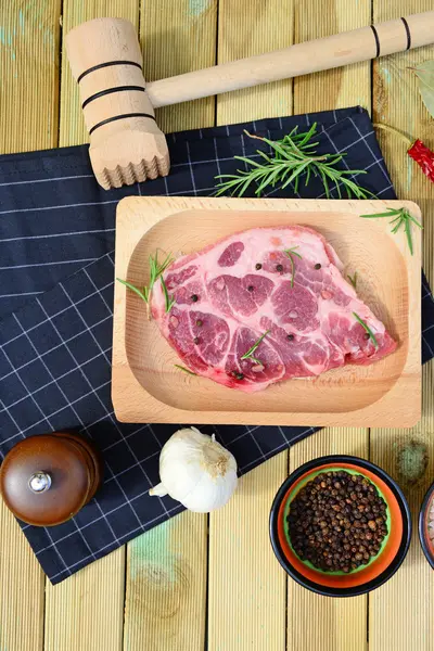 Viande de porc crue sur assiette en bois, moulin à poivre, romarin, épices et marteau sur fond en bois — Photo