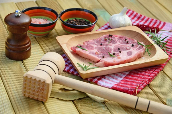 Rauw varkensvlees op houten plaat, peper molen, rozemarijn, specerijen en hamer op houten achtergrond — Stockfoto