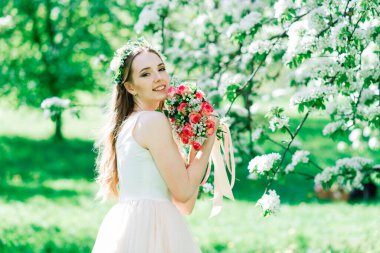 Beyaz gelinlikli gelin yeşil parkta bir buket tutuyor. Güneşli bir günde yaz düğünü..