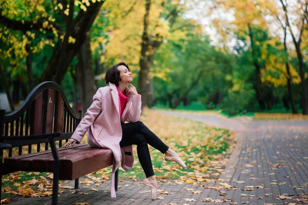 年轻美丽的芭蕾舞演员穿着漂亮的外套和尖尖的鞋子坐在长椅上 休息在秋天的公园里 — 图库照片
