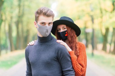 Genç kızıl saçlı kadın, sonbahar parkında genç bir adamla yürürken maske takıyor..