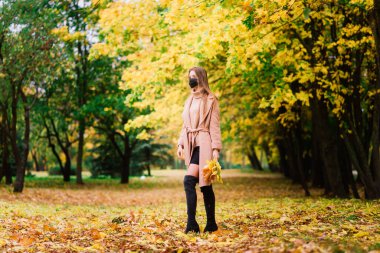 Parkta yürürken Corona virüsünden korunan genç bir kadın. Sonbahar arkaplanı.