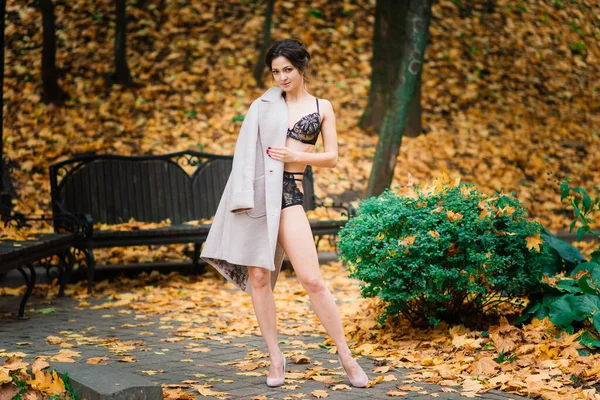 若い美しい黒髪の女性は ランジェリー姿と秋の公園で古典的なコートを身に着けています — ストック写真