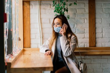 Maskeli genç Asyalı iş kadını telefon kullanıyor ve kahve dükkanının önünde oturuyor.