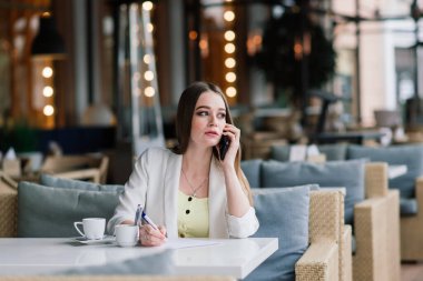 Beyaz ceketli genç bir kadın kafeteryada çalışıyor, kahve içiyor.
