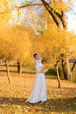 Uzun beyaz gelinlikli tatlı kız kırsal yolda poz veriyor ormandaki sonbahar ağaçları arasında altın saat atmosferinde.