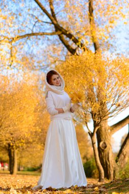 Uzun beyaz gelinlikli tatlı kız kırsal yolda poz veriyor ormandaki sonbahar ağaçları arasında altın saat atmosferinde.