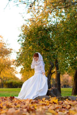 Uzun beyaz gelinlikli tatlı kız kırsal yolda poz veriyor ormandaki sonbahar ağaçları arasında altın saat atmosferinde.