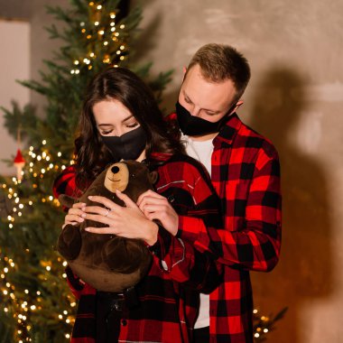 Tek kullanımlık tıbbi maskeli genç bir çift Noel ağacının altında. Karantina sırasında yeni yılla tanışma konsepti.