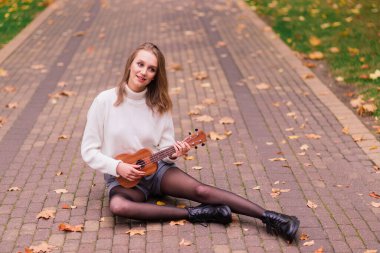Genç bir kadın parkta bankta oturmuş gitar çalıyor ve gitar çalıyor.