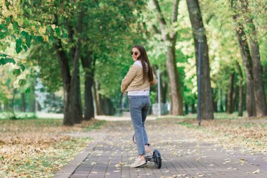 Sonbahar parkında elektrikli scooter kullanan genç bir kadın. Yeşil ulaşım, trafik sıkışıklığı.